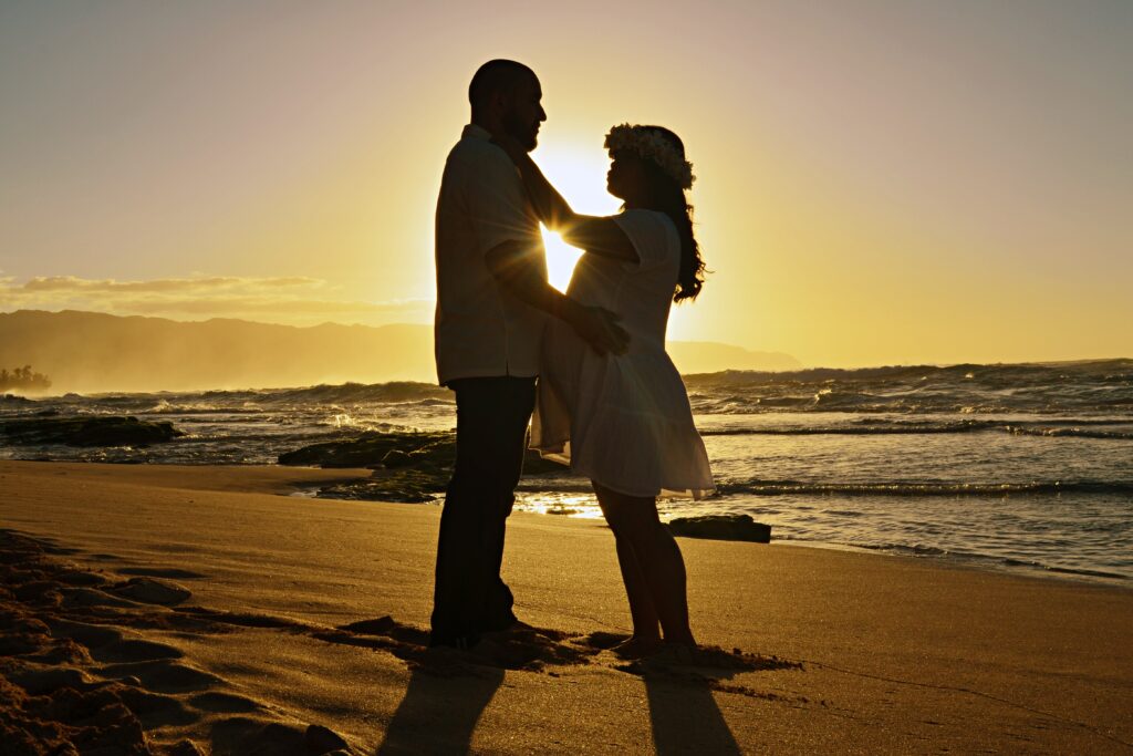 Pregnant couple standing on a beach in front of a sunset in Oahu, Hawaii in first maternity photography session