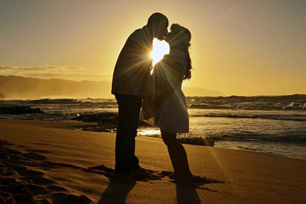 Pregnant couple standing on a beach in front of a sunset kissing in Oahu, Hawaii in first maternity photography session